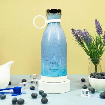 A white FreshJuice portable blender filled with a blue colored smoothie, surrounded by blueberries and a plant.