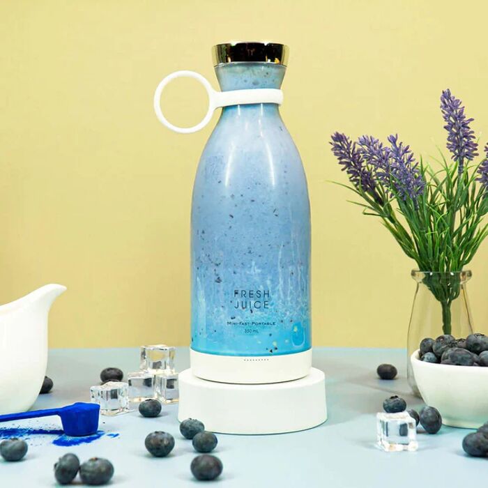 Un mixeur portable FreshJuice de couleur blanche rempli d'un smoothie de couleur bleue, entouré de bleuets et d'une plante avec des accessoires de cuisine tout autour