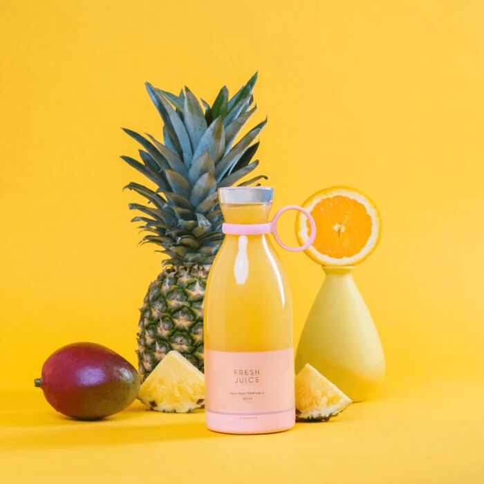 Un mini mixeur Fresh Juice rose, rempli de jus jaune et entouré d'un petit ananas et d'une demie orange sur fond jaune.