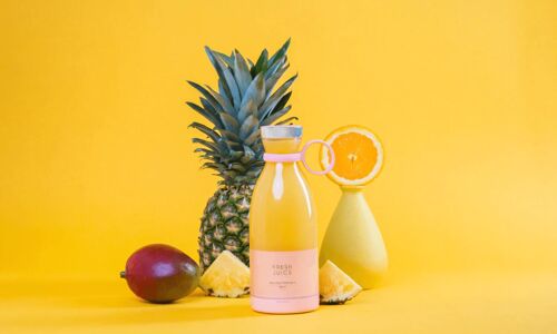 Un blender Fresh Juice de couleur jaune rempli d'un jus jaune et entouré d'ananas et d'oranges sur un fond jaune.