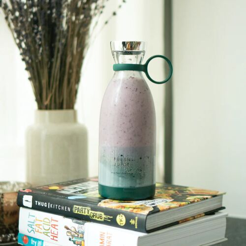 Ein grüner Fresh Juice Mixer, gefüllt mit einem rosa und weißen Smoothie, der auf zwei Büchern vor einer Pflanze in einer Vase sitzt.