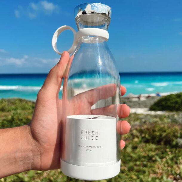 Der tragbare elektrische Mixer Fresh Juice vor dem Meer mit einer Hand, die ihn hält, um seine Tragbarkeit zu demonstrieren.